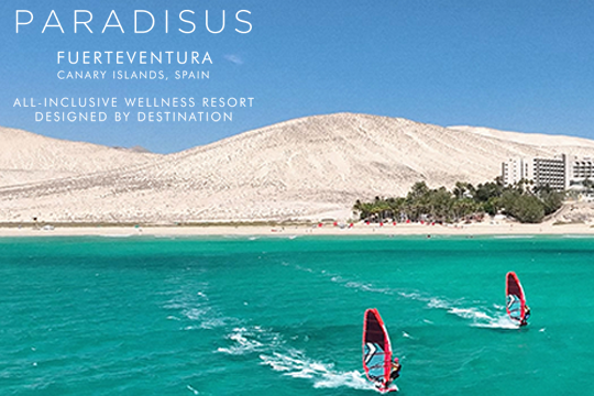 Zwei Surfer im Vordergrund, hinten Sanddünnen und rechts ein Hotel